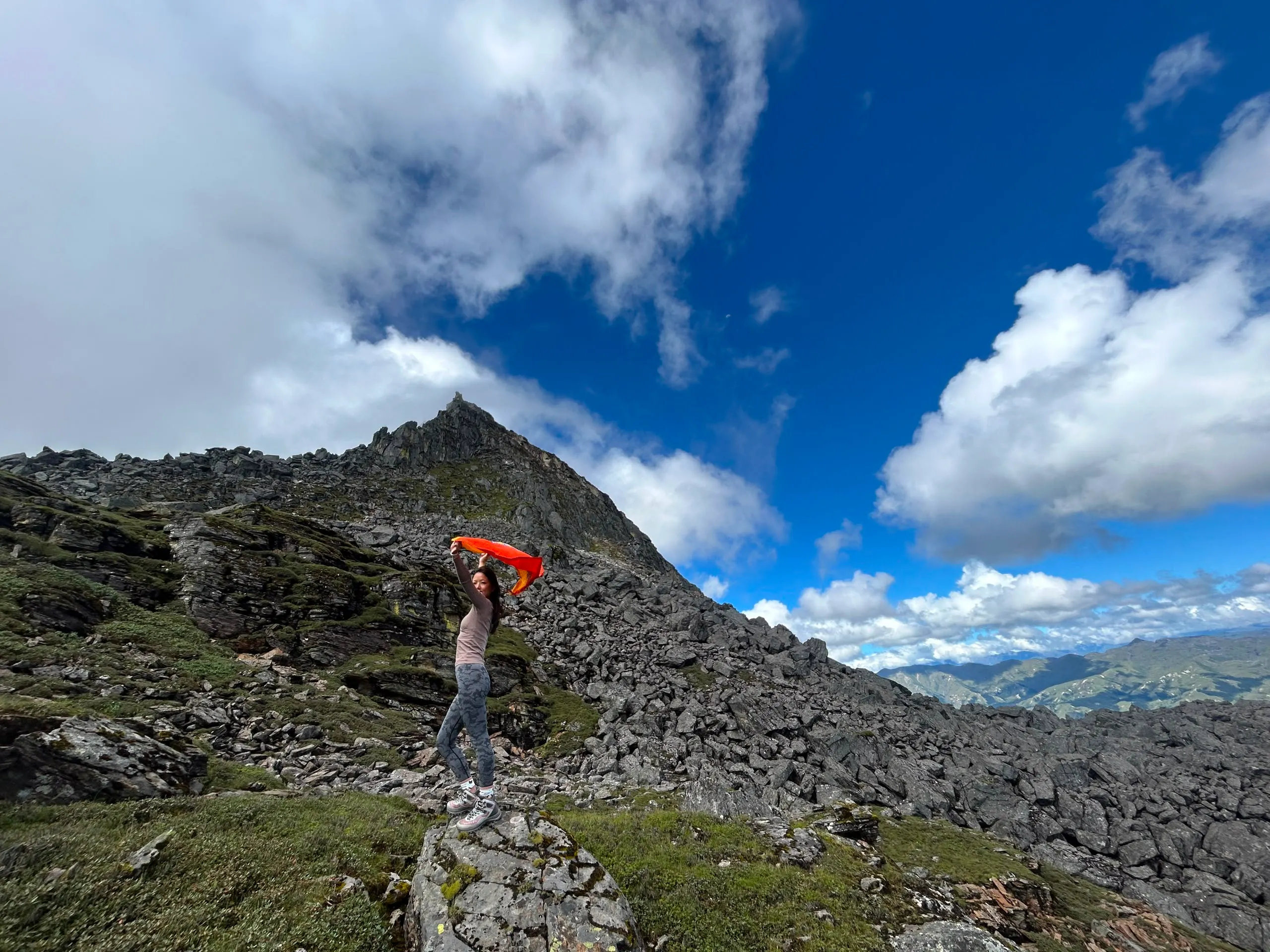 A trekker on a high rocky Bhutanese ridge holding a scarf to the wind below a rugged peak