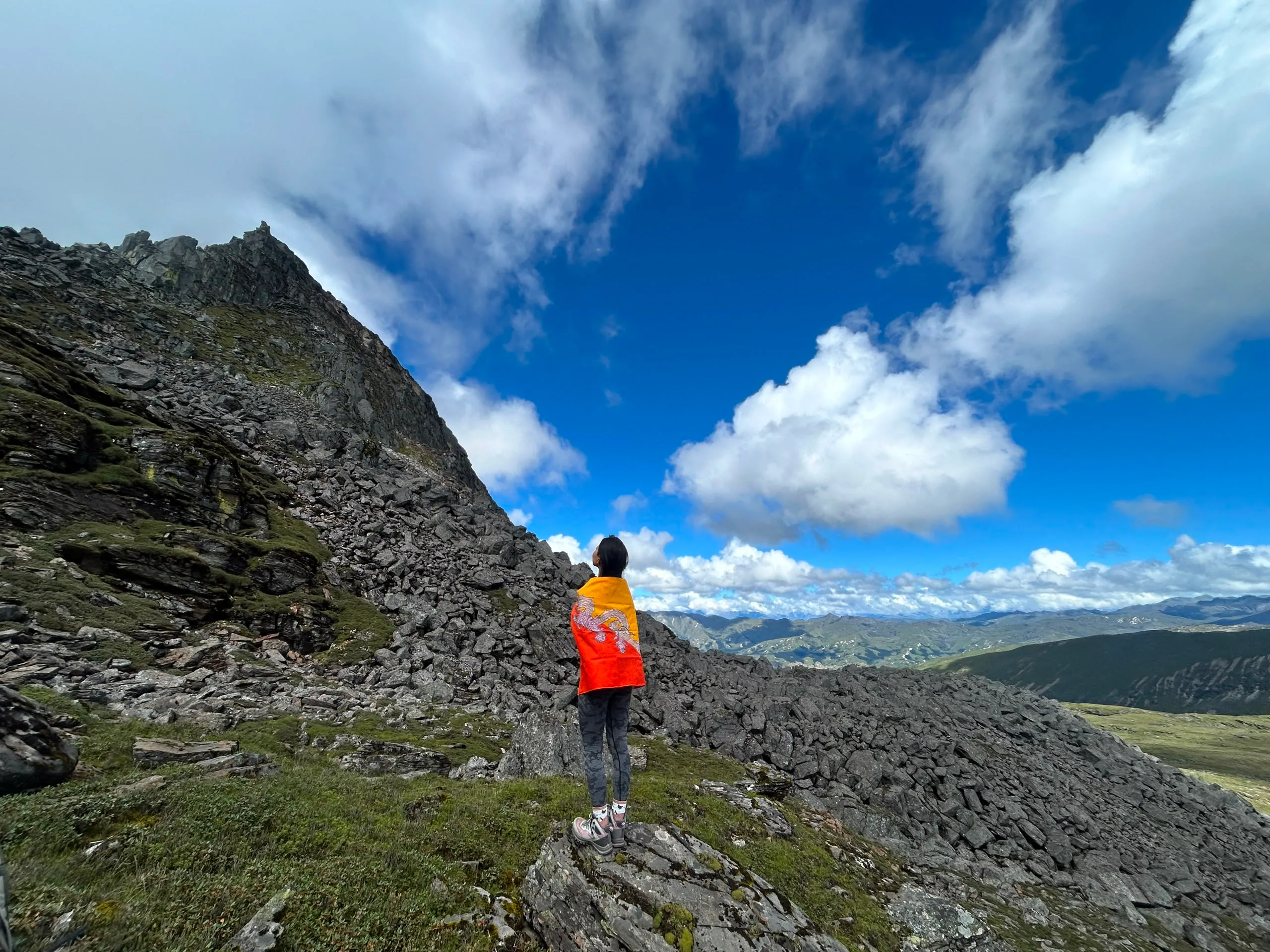 A traveller draped in the Bhutan flag on a ridge, looking over the mountains