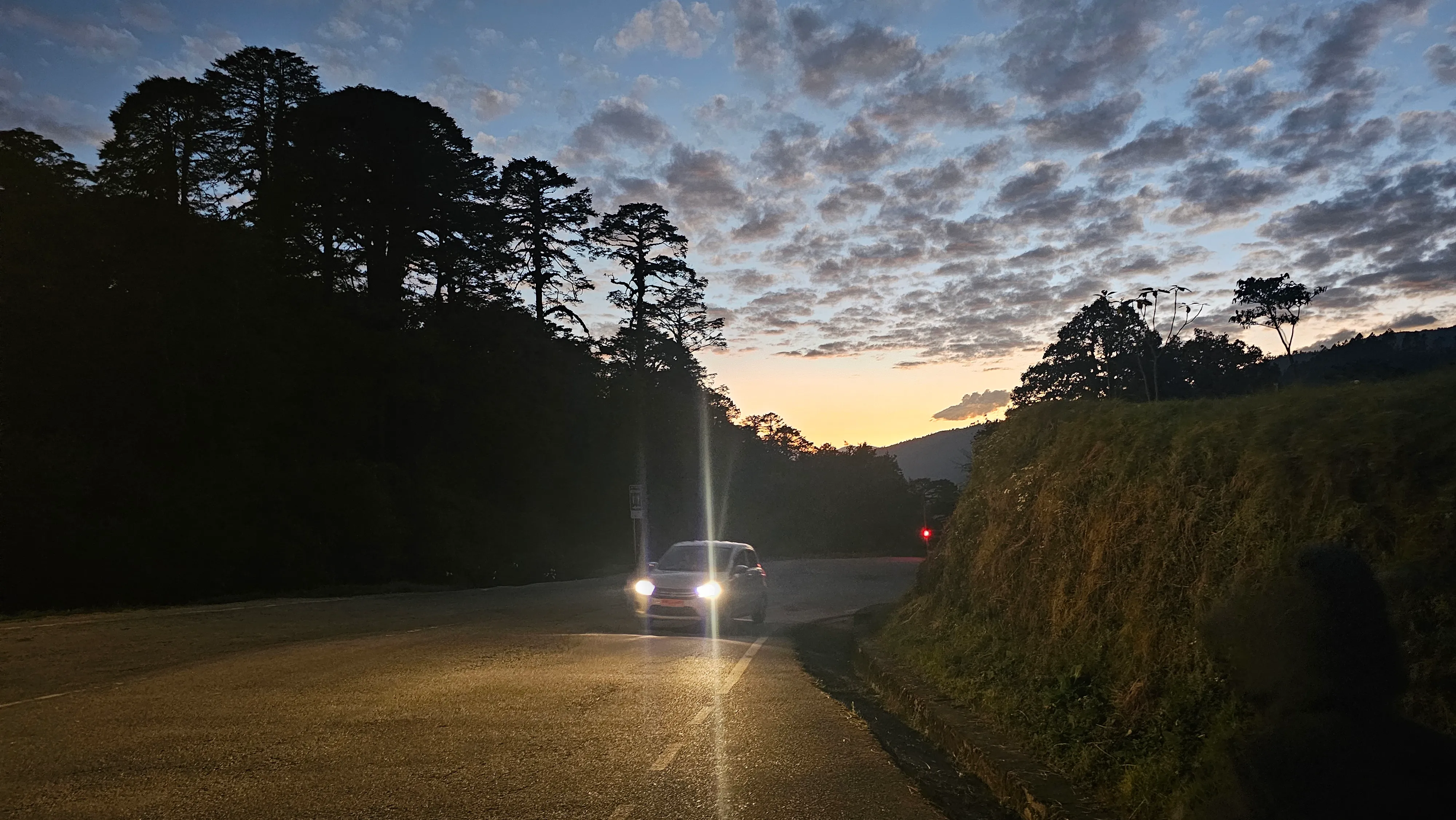 A car on a Bhutanese mountain road at dusk with silhouetted pines and a glowing sky
