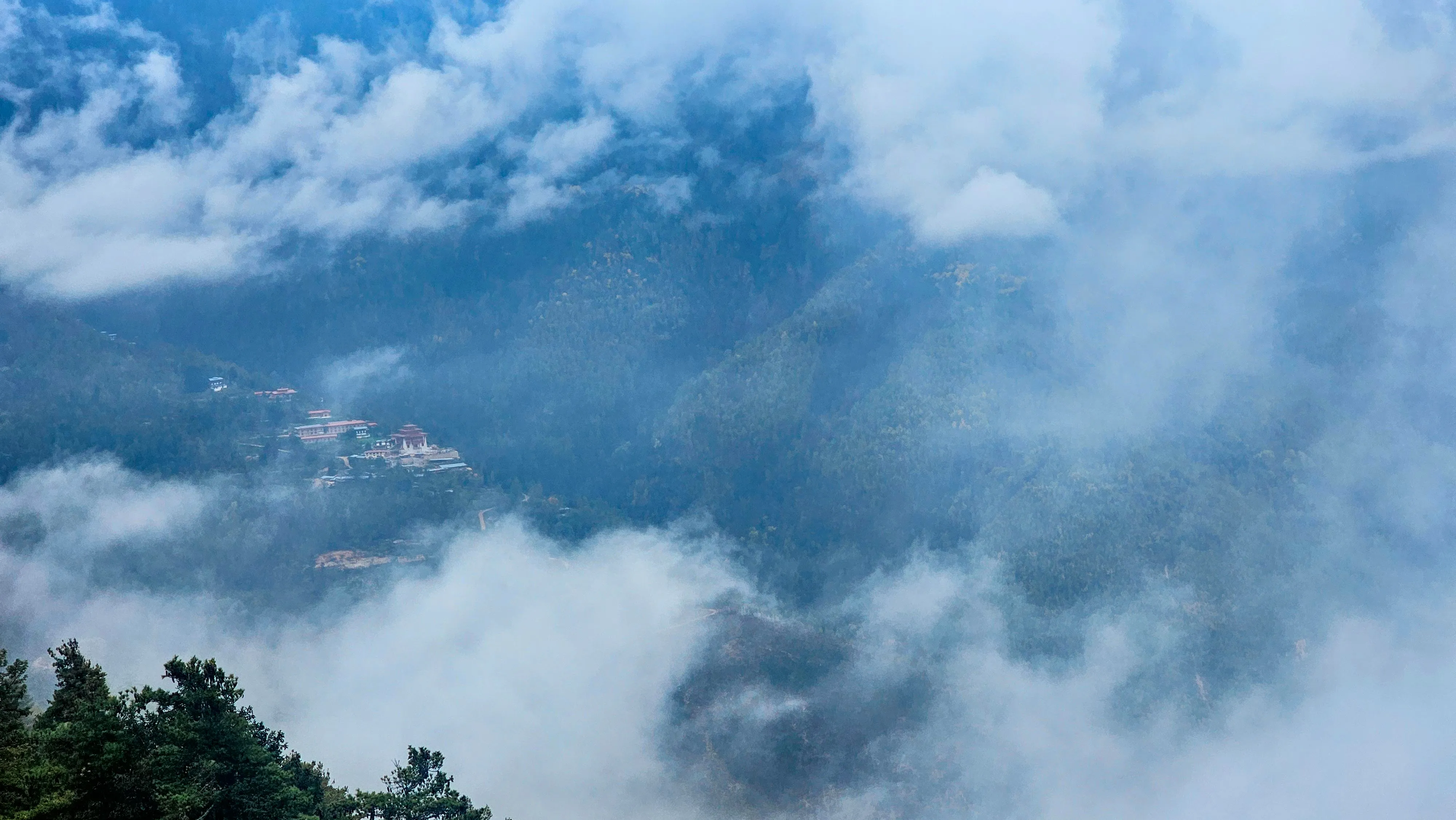 A dzong on a forested Bhutanese hillside with clouds drifting through the valley