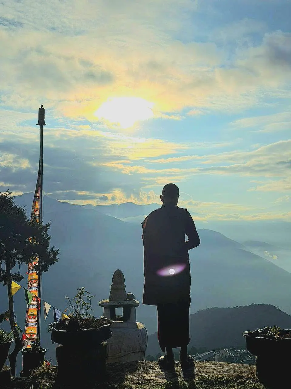 A monk silhouetted on a ridge at sunset, looking out over misty Bhutanese mountains and prayer flags