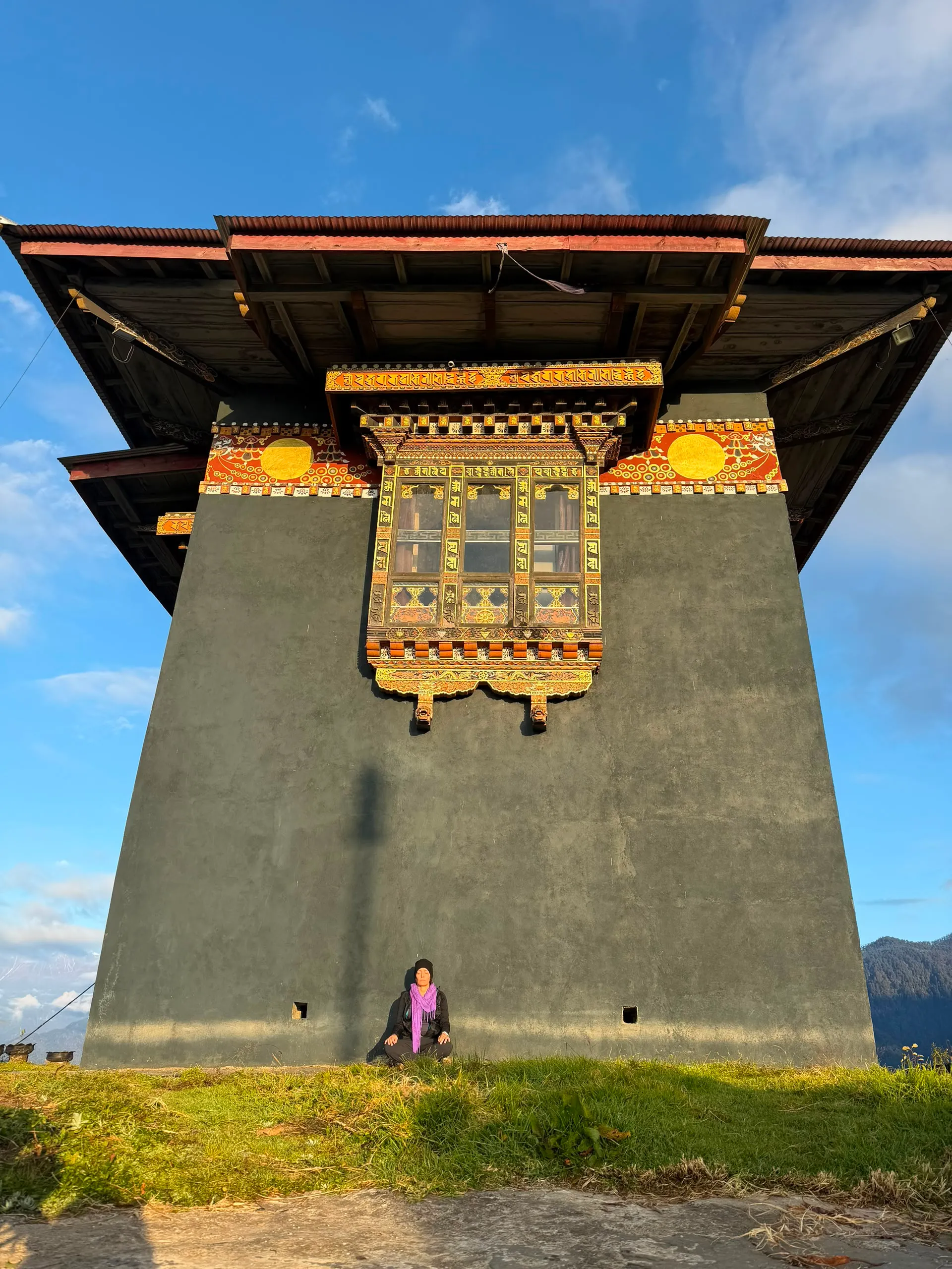 A traveller meditating on the grass below a tall painted Bhutanese temple window