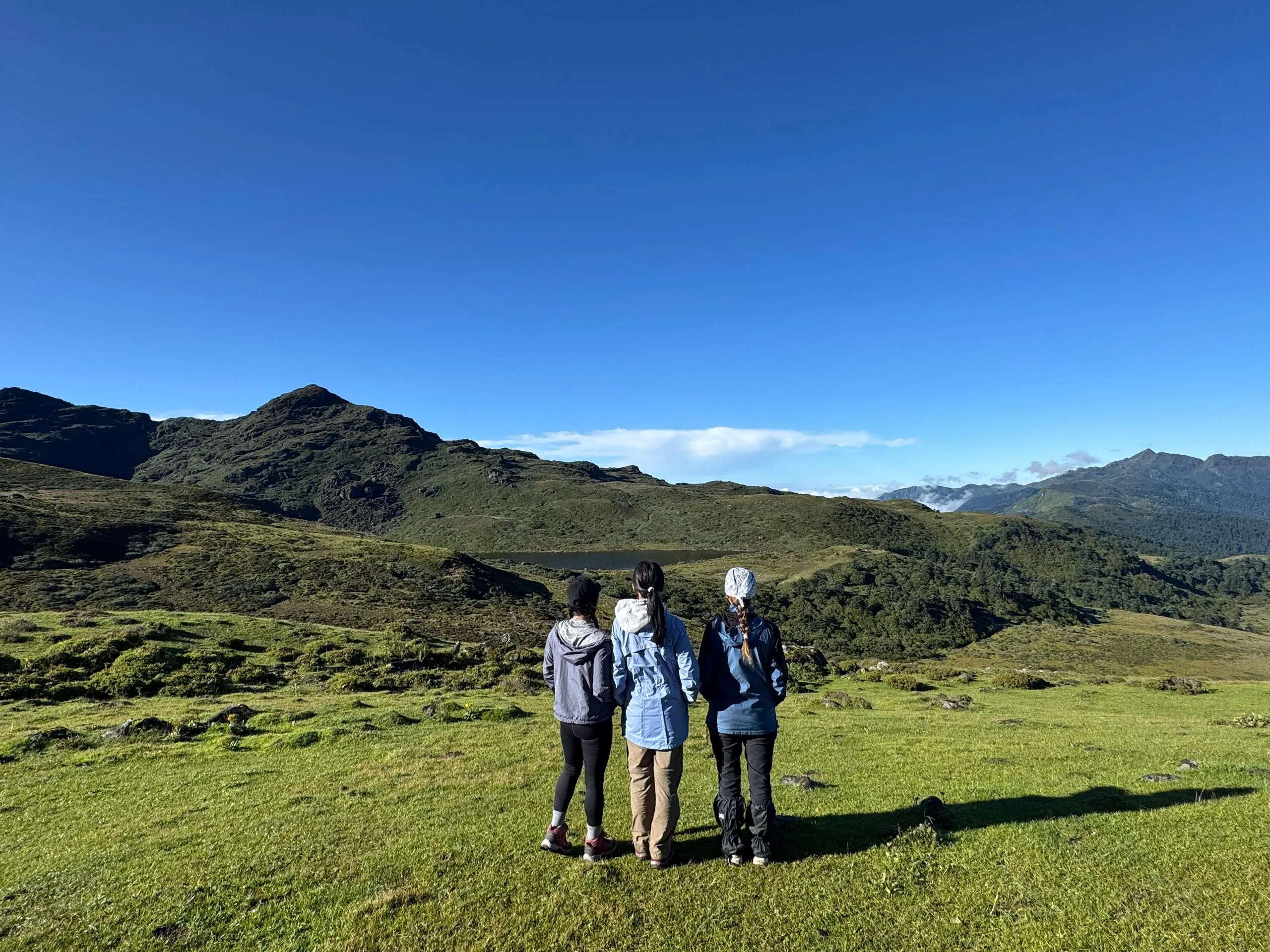 Three trekkers looking out over a high green valley and mountain lake in Bhutan